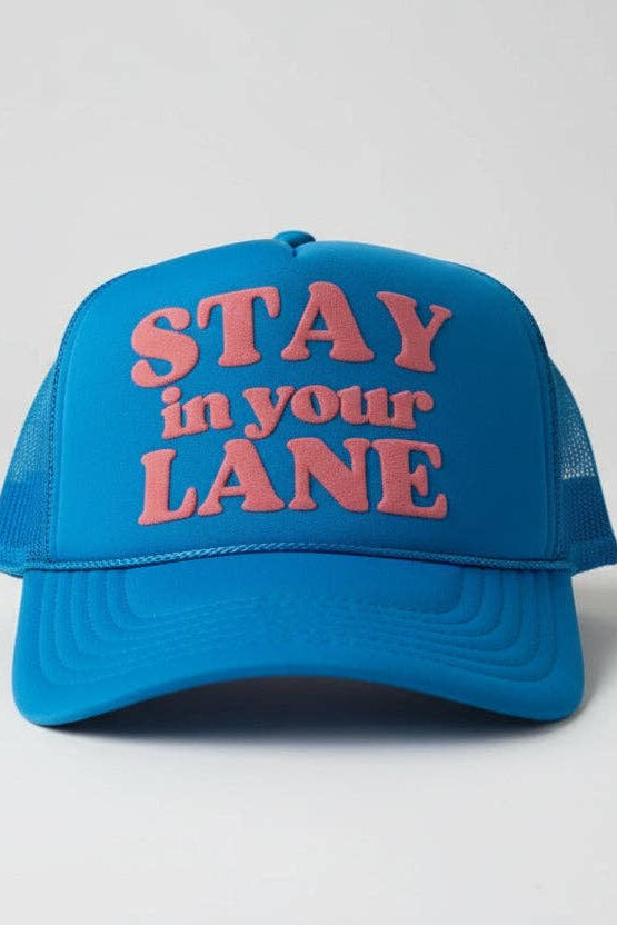A bright blue trucker hat with pink embroidered text reading 'STAY in your LANE' on the front, shown against a light gray studio background.