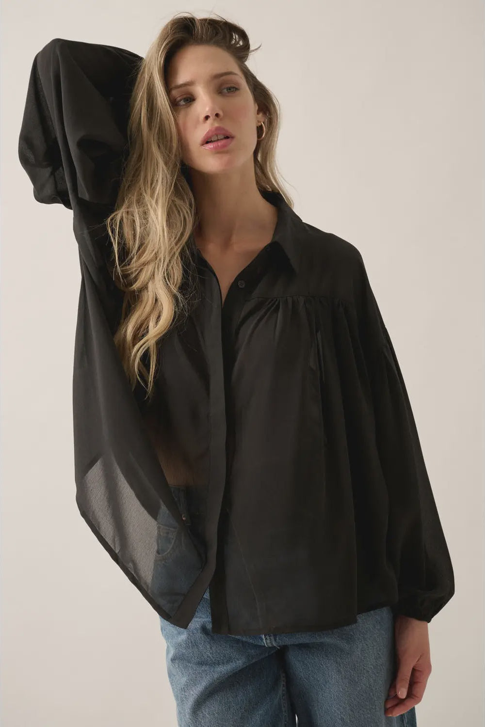 Woman wearing a black sheer blouse over a dark top with blue jeans on a plain background
