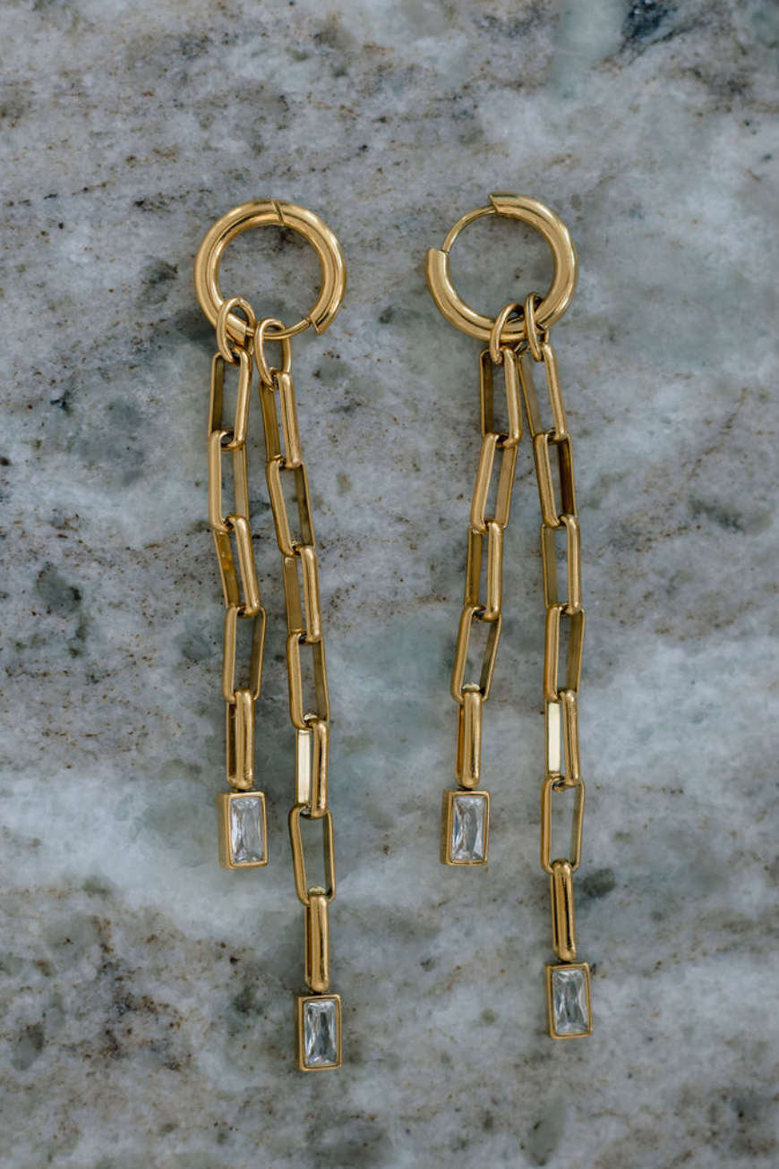 A pair of gold dangle earrings with rectangular chain links and clear baguette stones, displayed on a gray and white marbled surface.