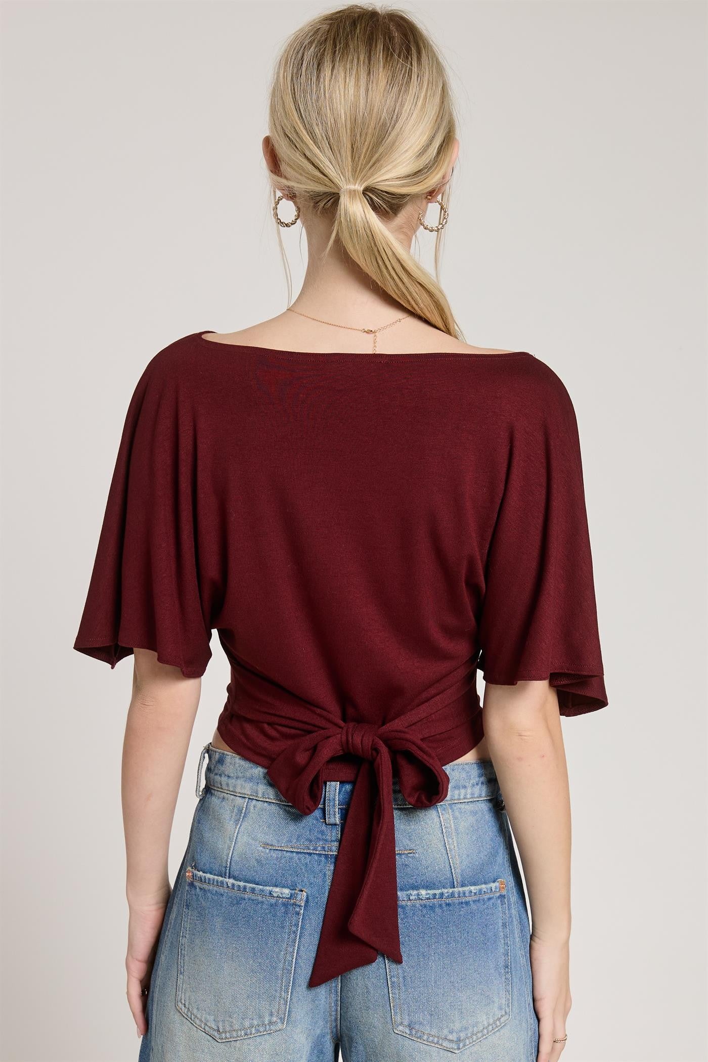 Woman wearing a burgundy top with a tie detail and blue jeans on a plain background