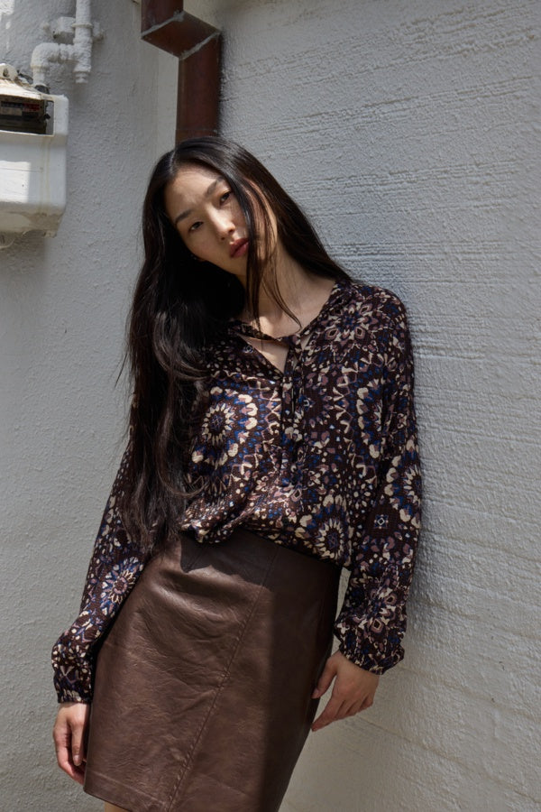 Woman wearing a patterned blouse and brown skirt against a gray wall.