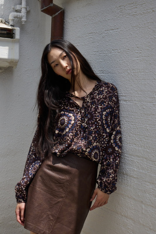 Woman wearing a patterned blouse and brown skirt against a gray wall.