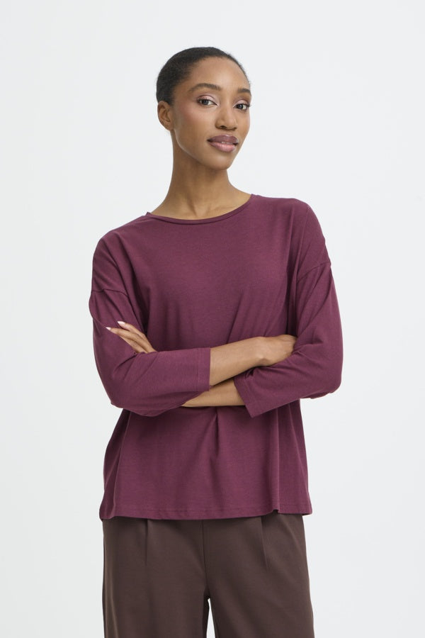 A woman modeling a long-sleeve crewneck top in a muted plum or burgundy shade, paired with brown pants, against a clean light gray studio background.