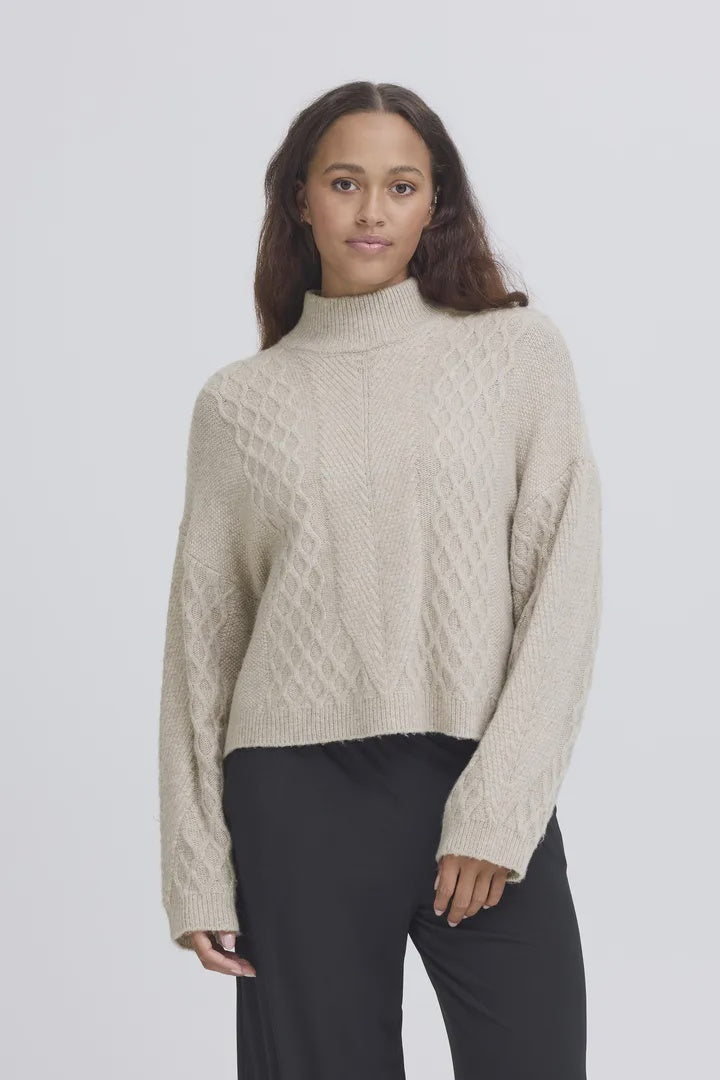 Woman wearing a beige cable knit sweater and black pants on a light gray background