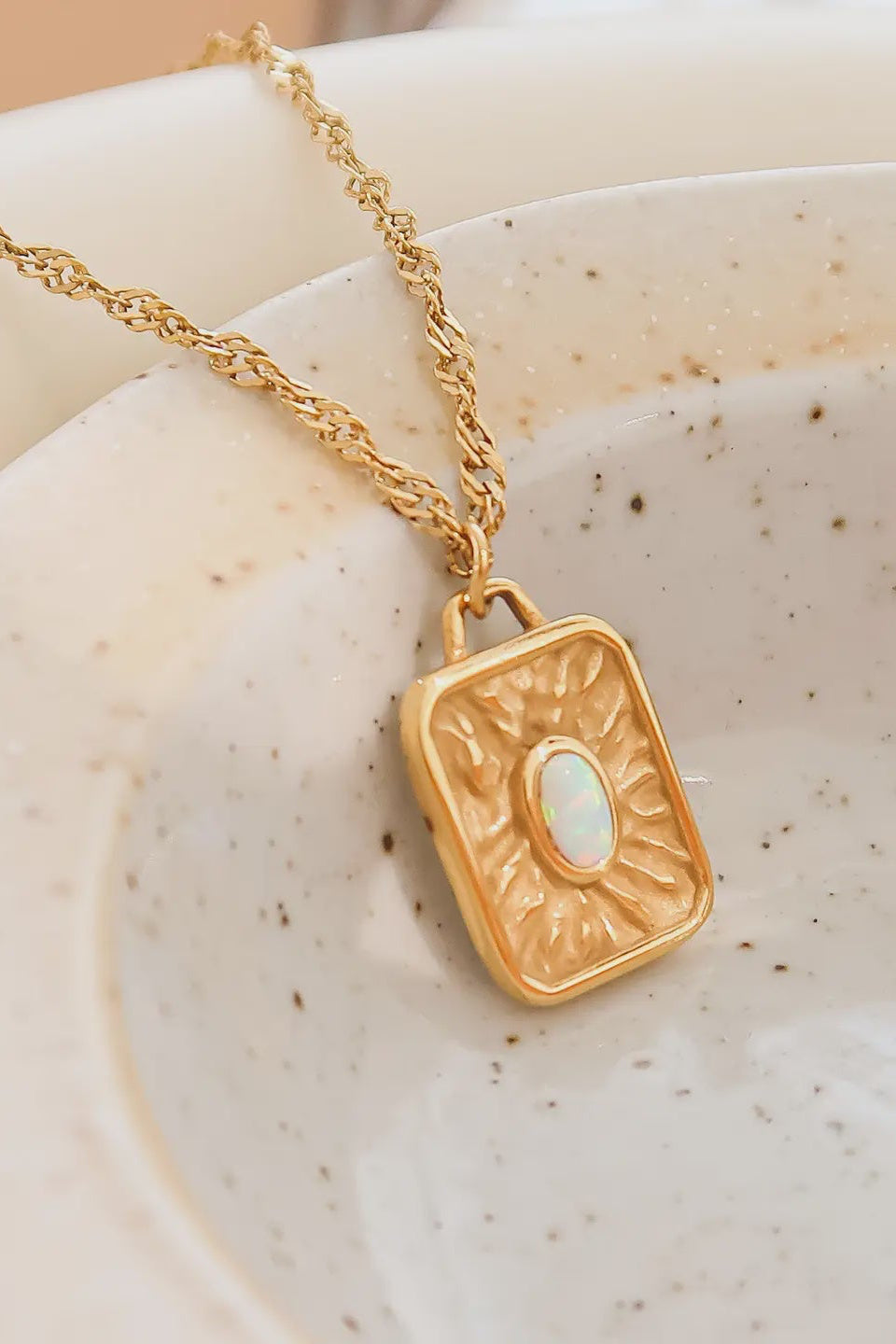 A gold-plated stainless steel pendant necklace with a rectangular sun design, displayed on a ceramic plate.