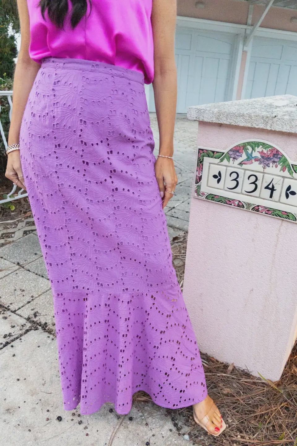 A person wearing a bright pink satin top and a lavender eyelet lace maxi skirt, standing outdoors next to a mailbox.