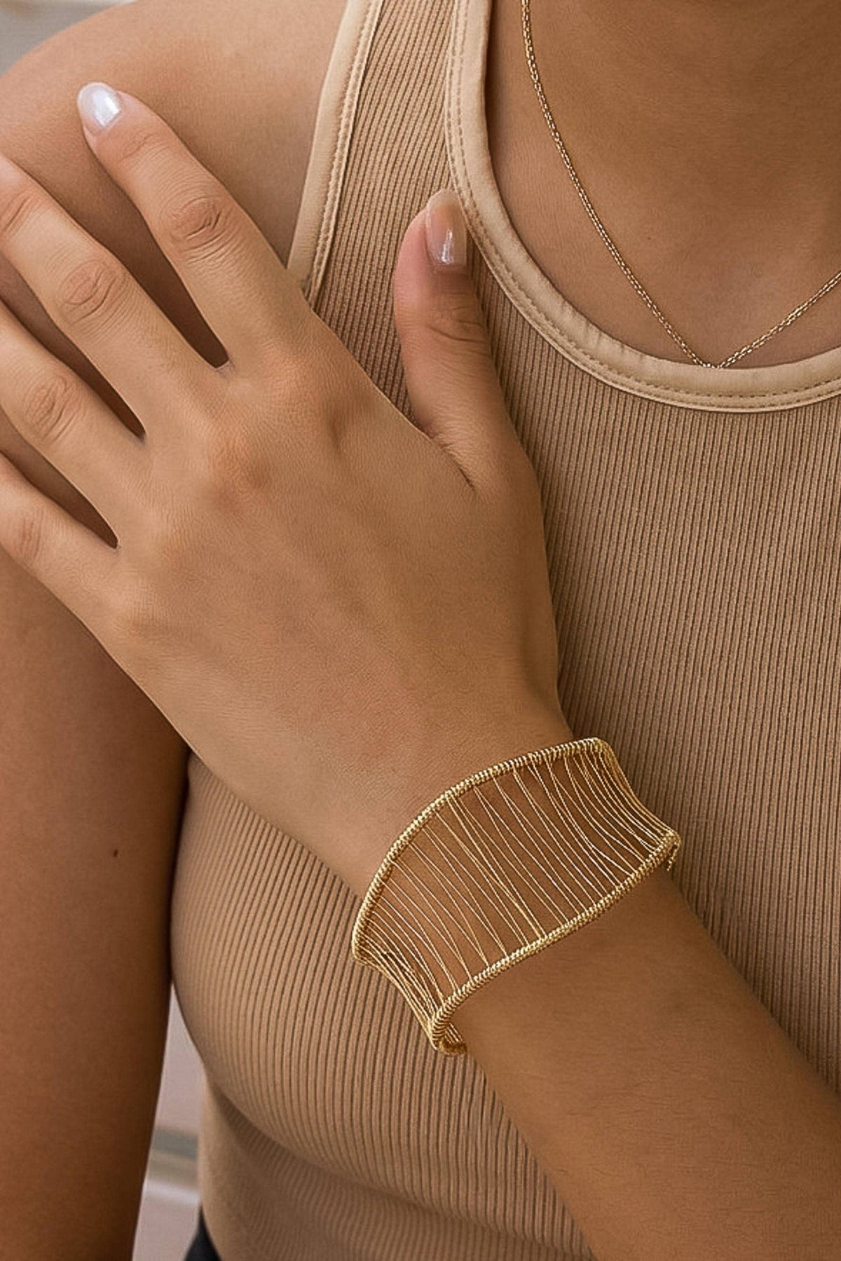 A close-up of a person wearing a wide, open-weave gold cuff bracelet on their wrist, paired with a beige ribbed tank top and a delicate gold necklace.