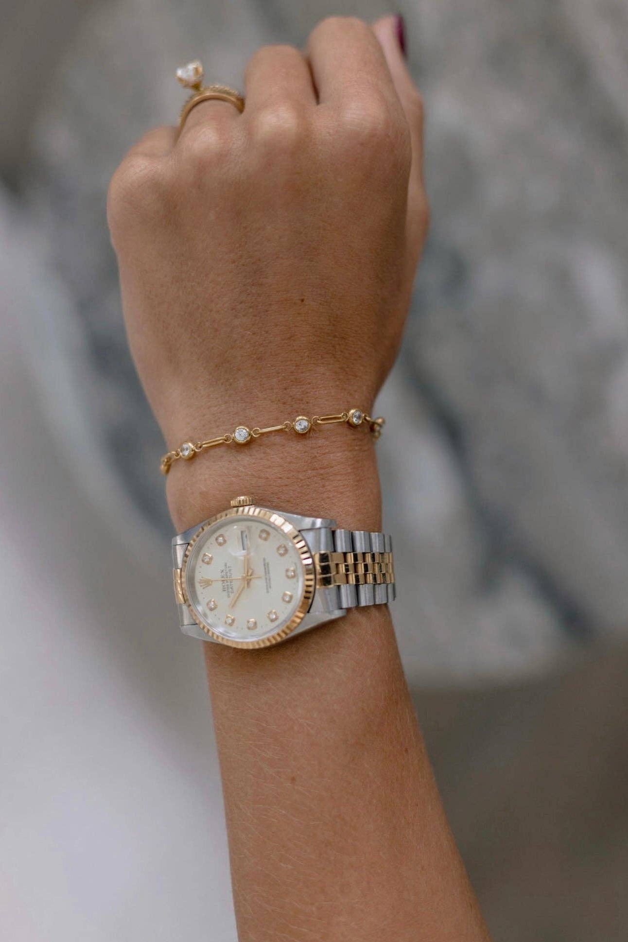 A close-up of a person's wrist wearing a two-tone gold and silver watch with a white dial, along with a delicate gold bracelet and ring, against a soft gray background.