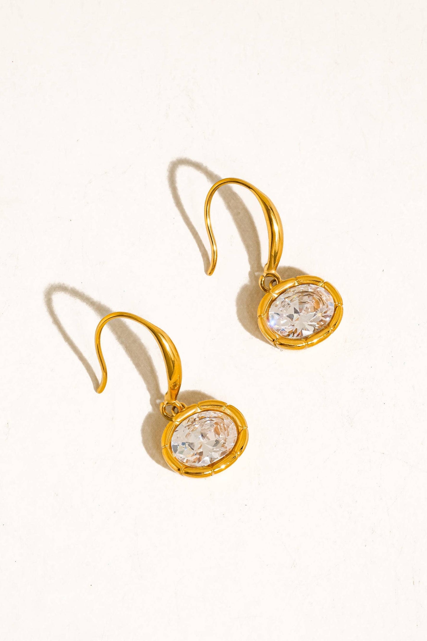 A pair of gold drop earrings with round clear faceted gemstones in bezel settings, displayed on a warm off-white background.
