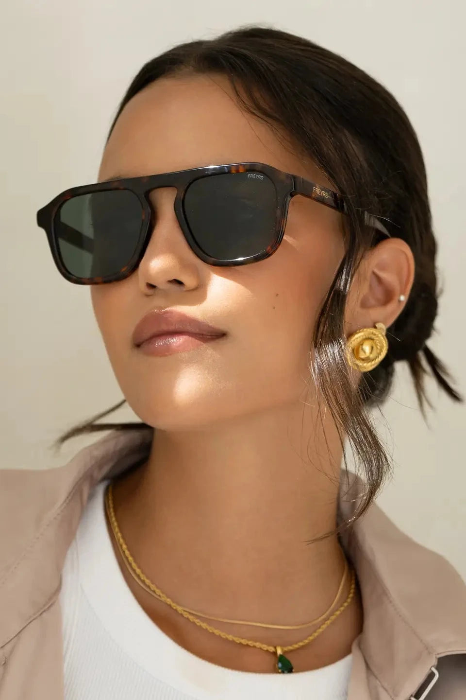Woman wearing sunglasses and gold jewelry against a neutral background