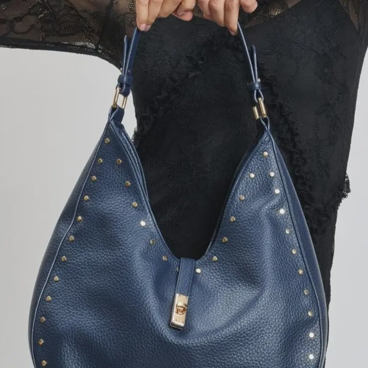 Blue leather handbag with studs held by a person wearing black pants.