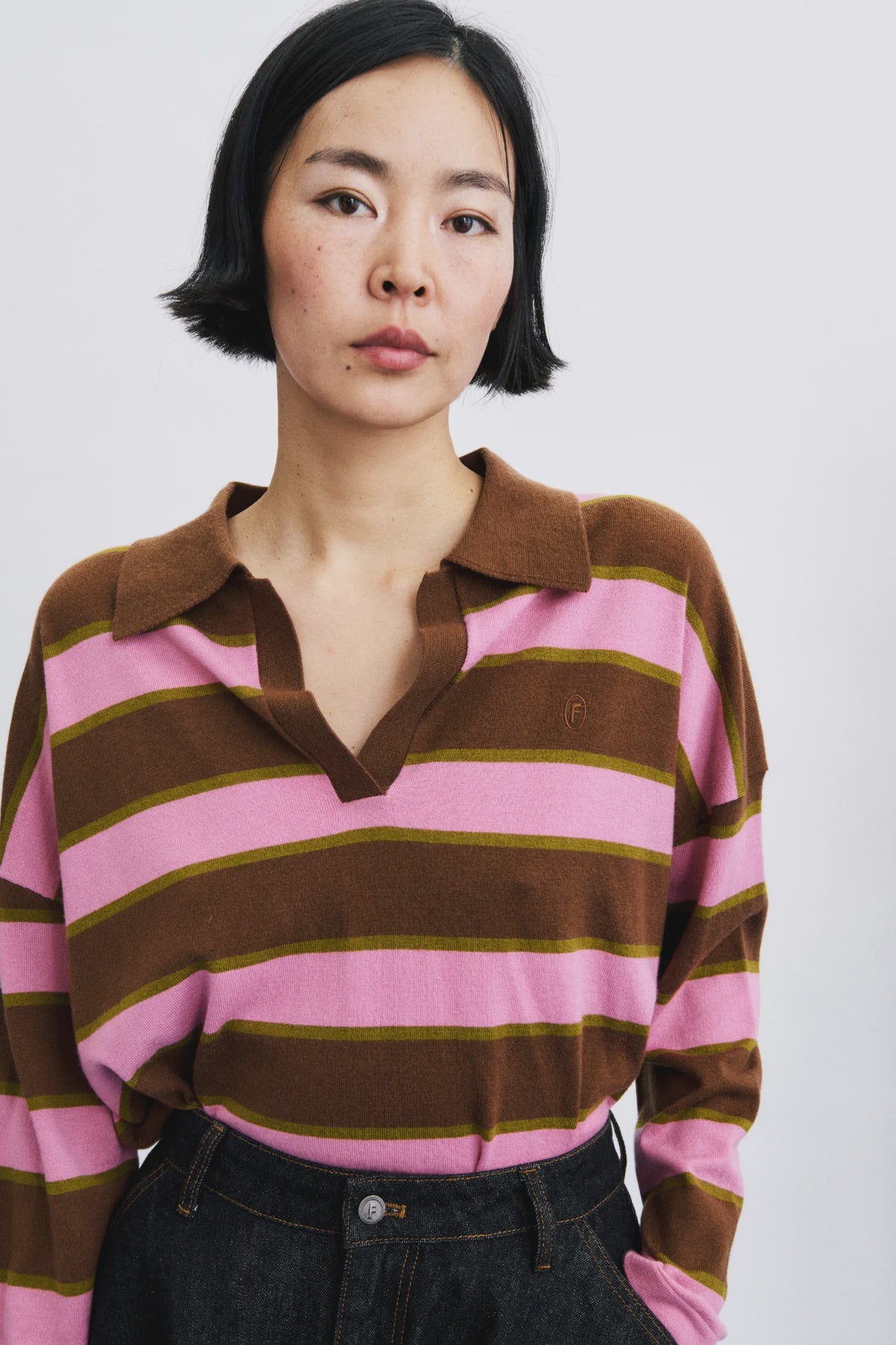 A woman wearing a pink and brown striped long-sleeve polo shirt with dark denim jeans, photographed against a light gray studio background.