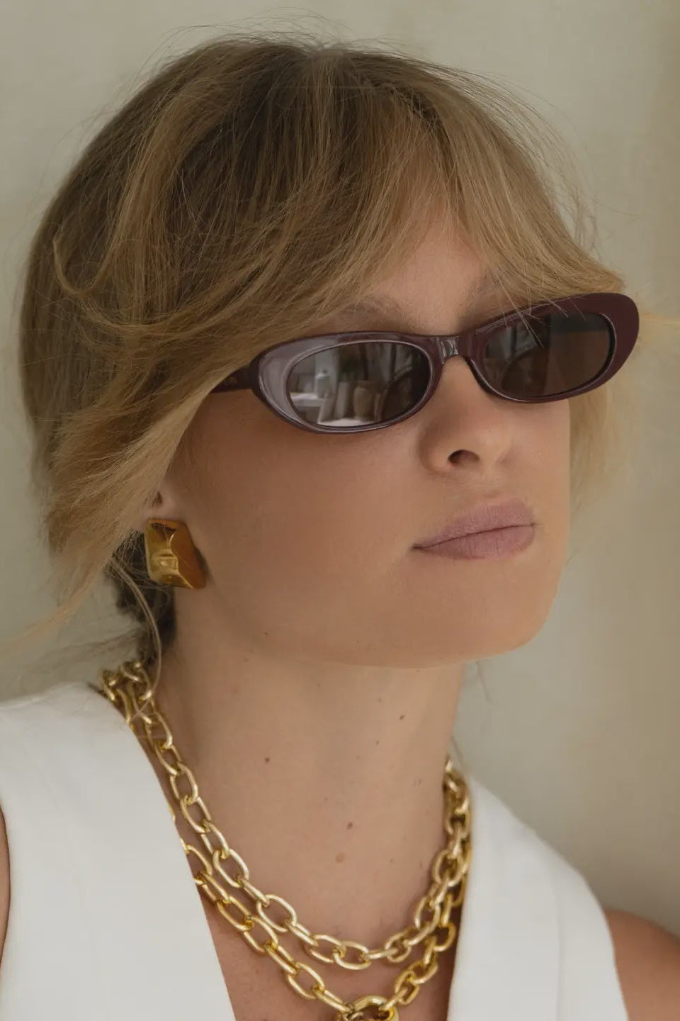 Woman wearing sunglasses and gold jewelry against a neutral background