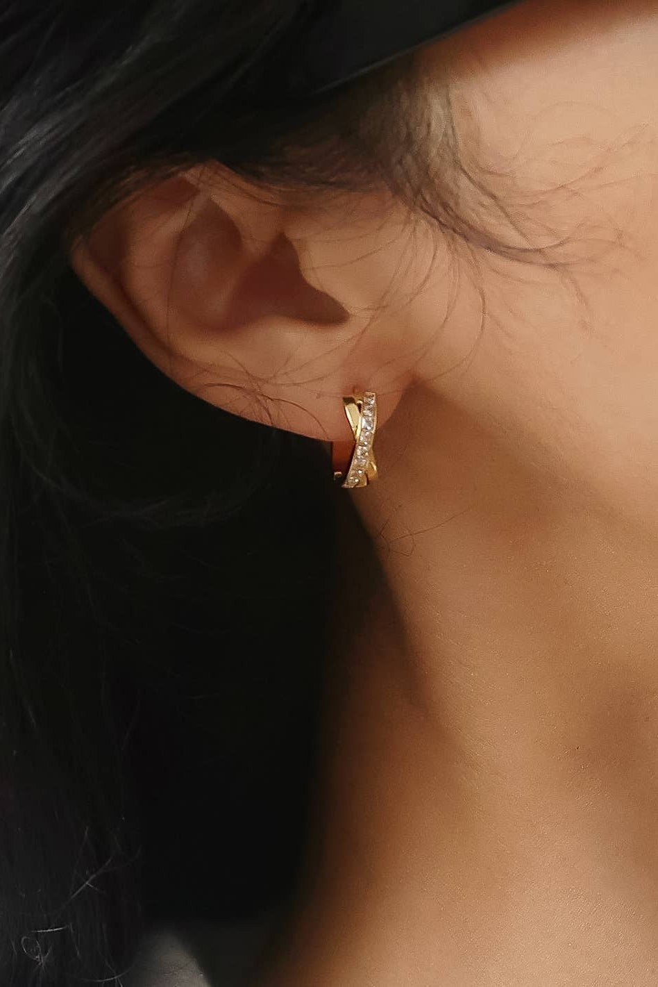 A close-up of a person's ear wearing a small gold huggie hoop earring with clear stones, against dark hair and warm skin tones.