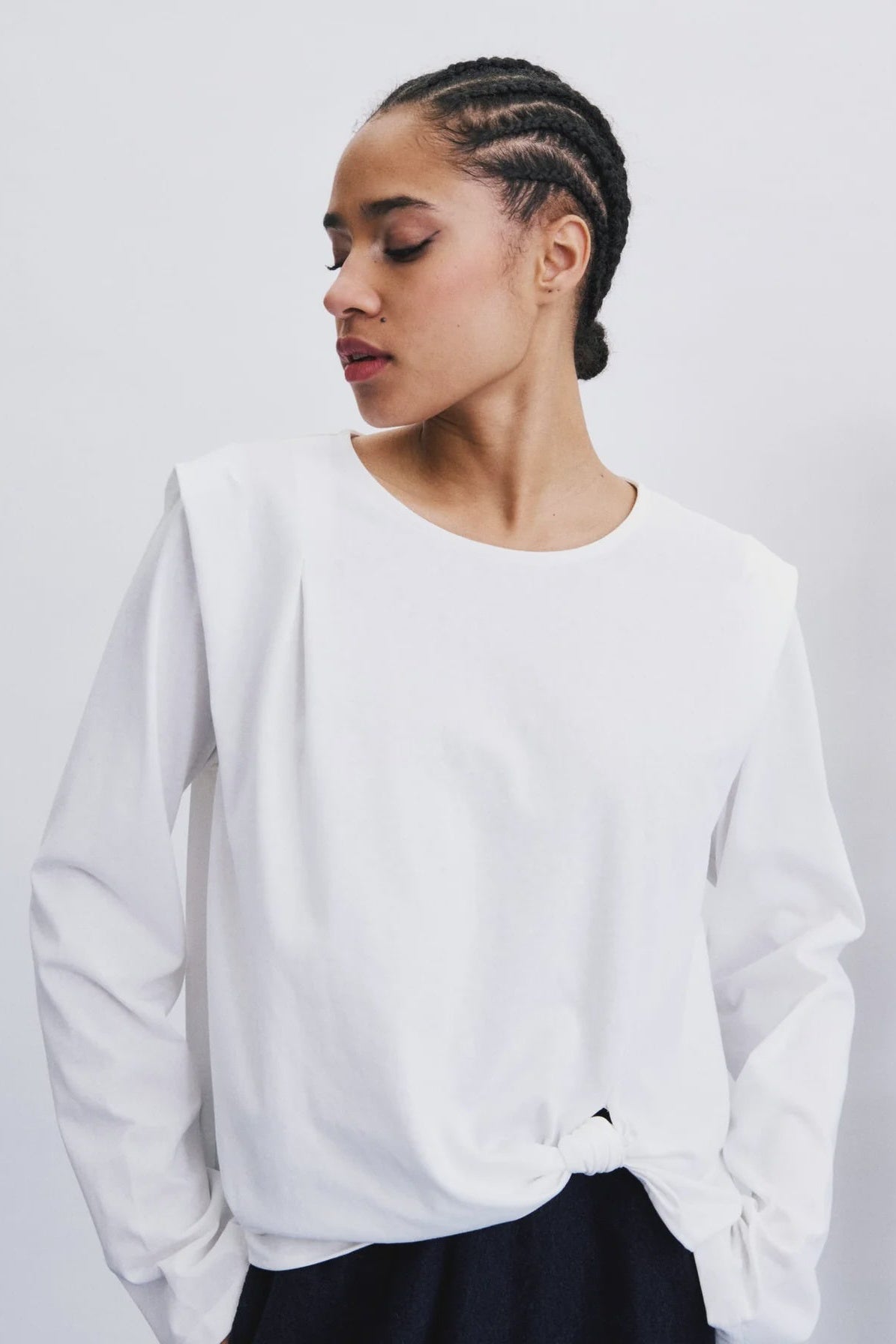 A woman with braided hair wearing a white long-sleeve top with a crew neck and a side knot detail, paired with a dark skirt, against a plain light gray background.