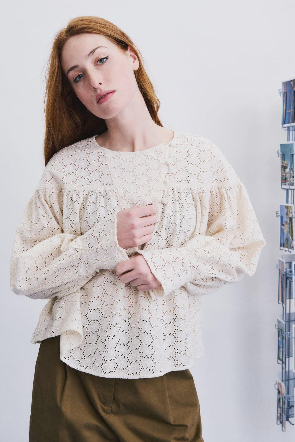 A woman wearing a cream-colored, long-sleeved lace blouse with a gathered ruffle at the neckline, paired with olive green pants, standing against a white wall next to a wall-mounted photo rack.