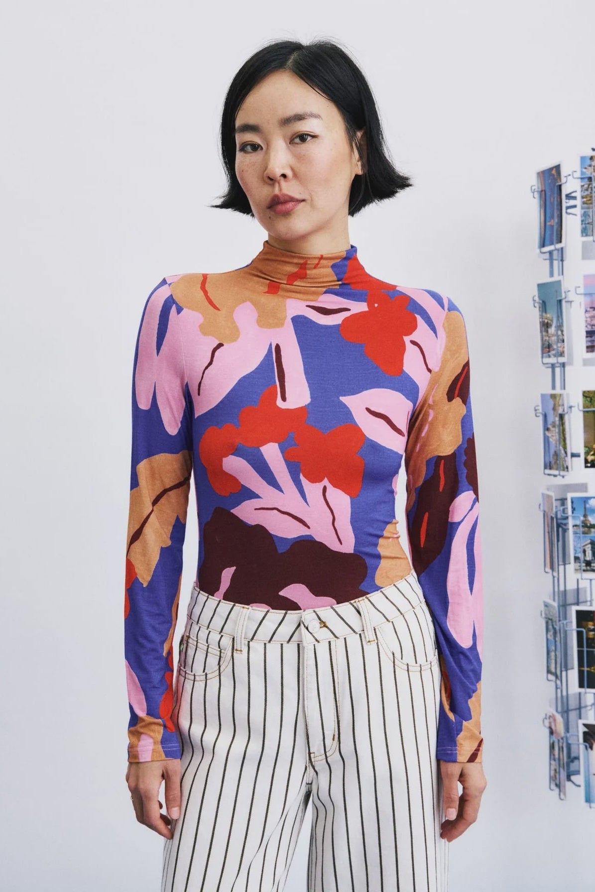 A woman modeling a long-sleeve mock neck top with a bold abstract floral pattern in pink, purple, red, and brown, paired with white and black pinstriped trousers, standing against a light gray wall.