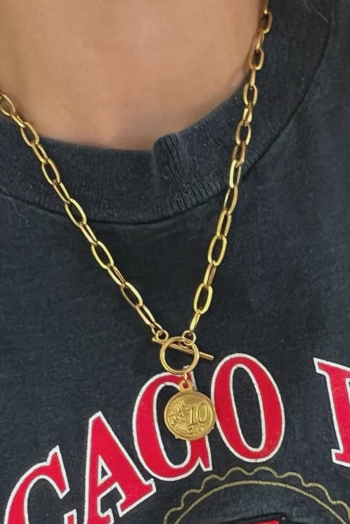 Gold chain necklace with a pendant on a person wearing a dark shirt with red text.