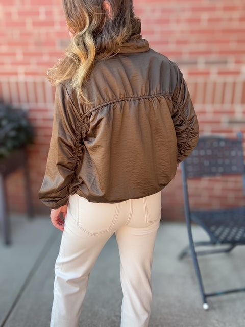 Person wearing a brown jacket and white pants standing in front of a brick wall.