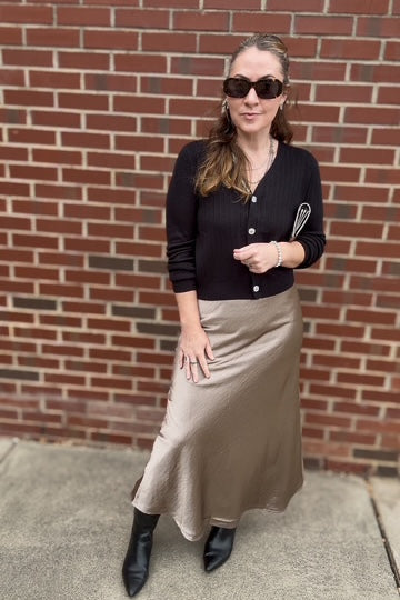 Woman wearing a black cardigan, beige skirt, and sunglasses standing in front of a brick wall.