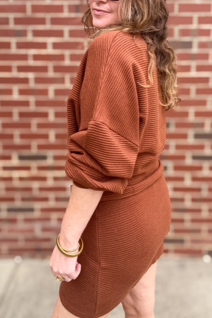 Person wearing a brown ribbed dress against a brick wall.