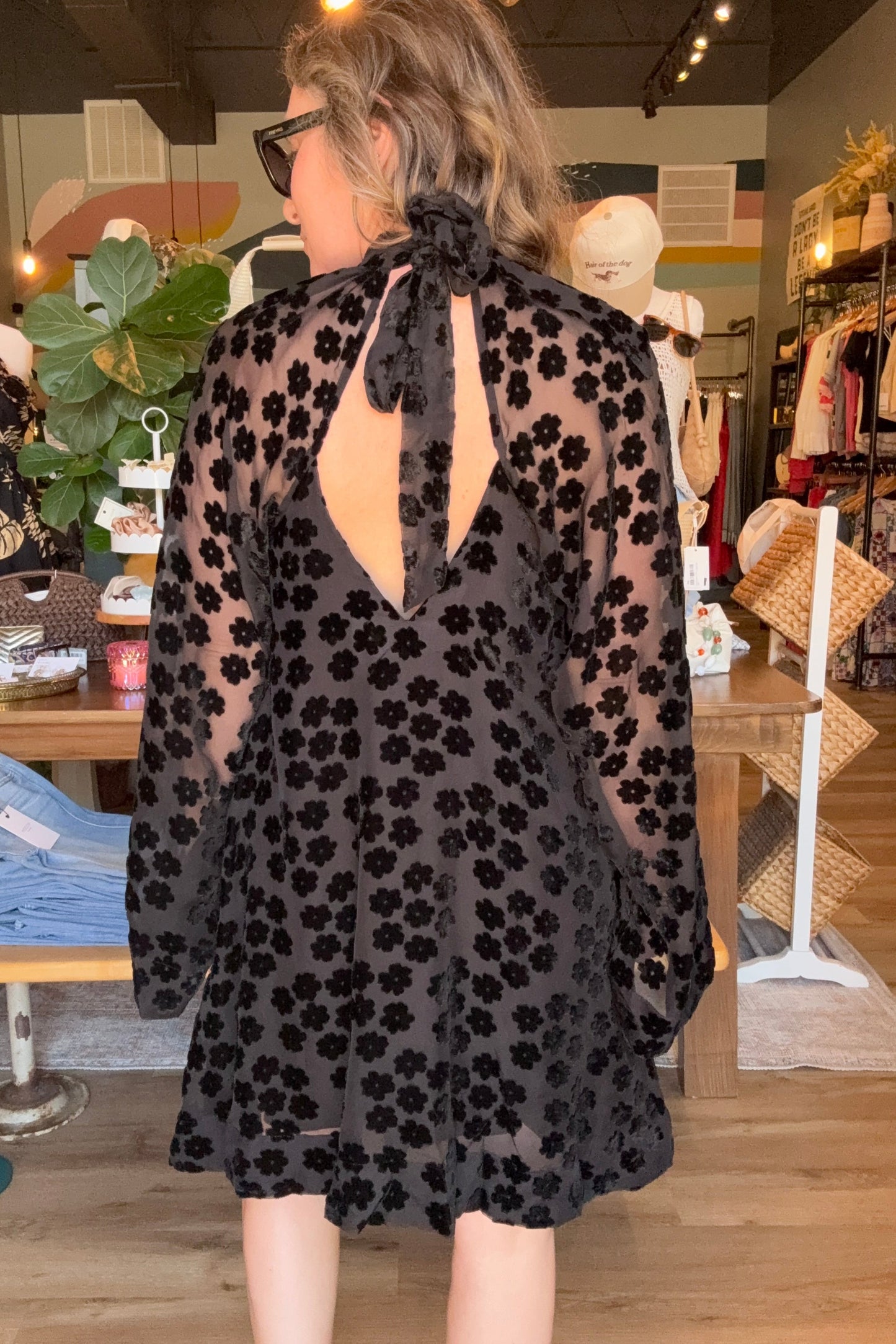 A woman wearing a sheer black long-sleeve dress with a black floral pattern and a large back tie, photographed from behind inside a clothing boutique.
