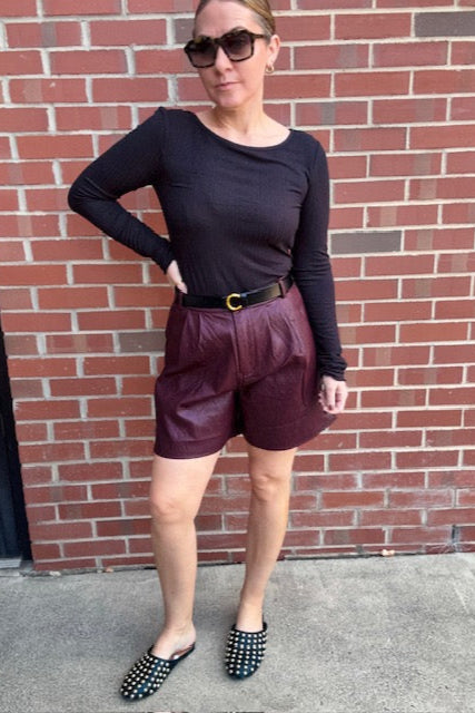 Woman wearing a black long-sleeve top and maroon shorts standing against a brick wall.