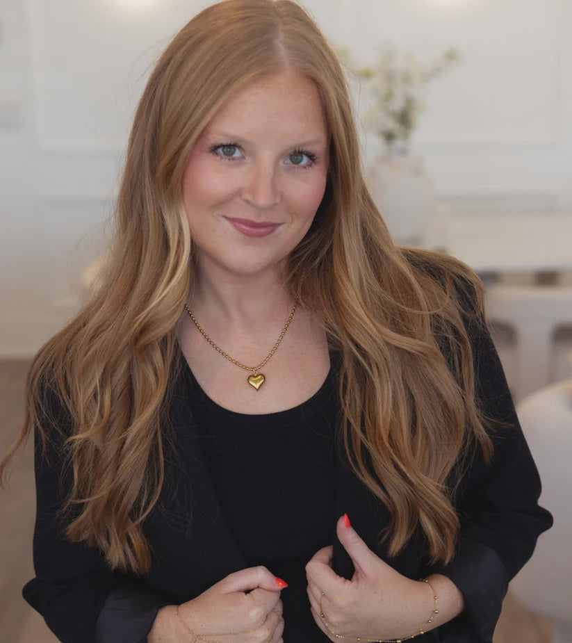 Woman with long blonde hair wearing a black top and gold necklace indoors.
