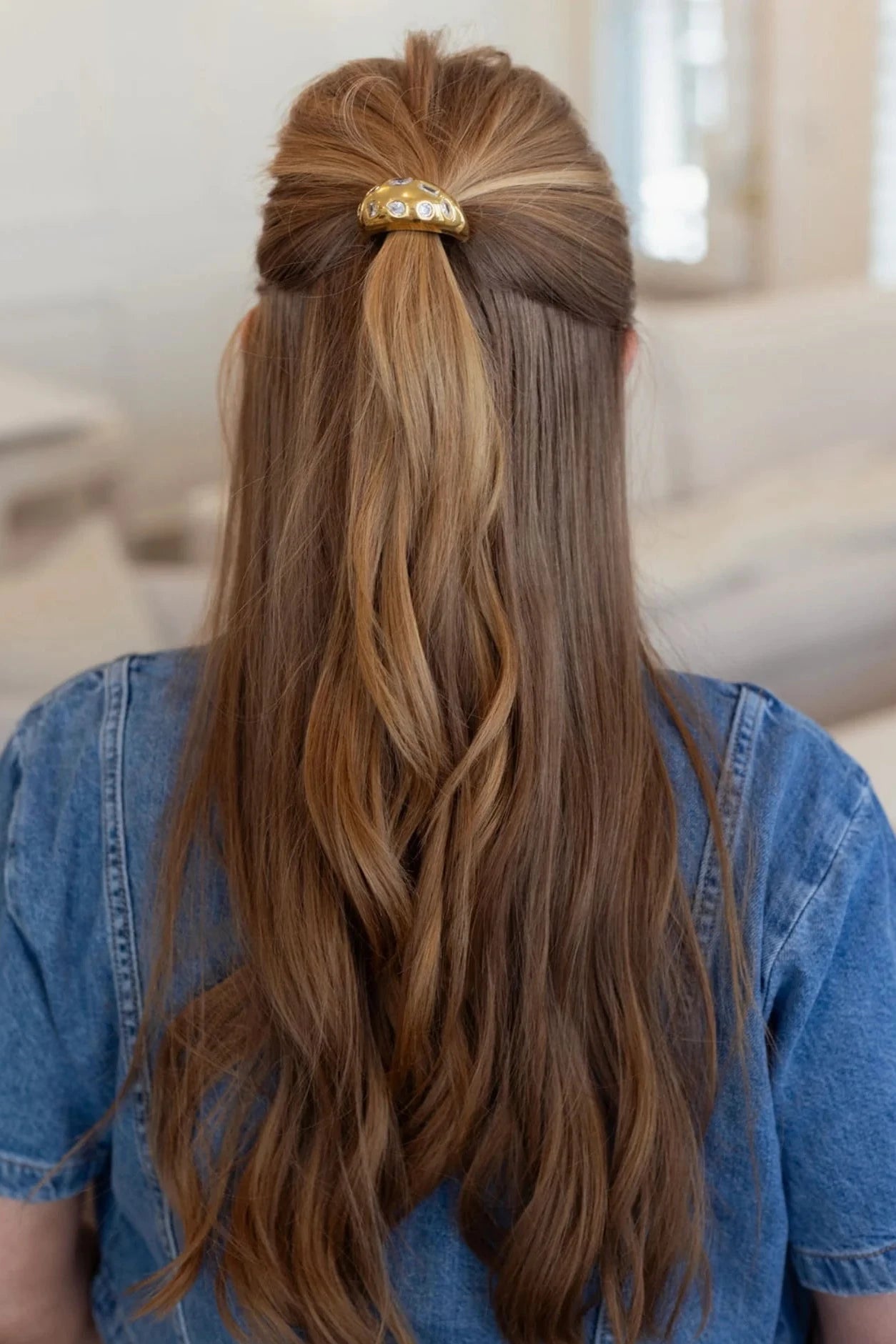 Person with long, wavy hair styled in a half-up, half-down look with a gold hair accessory, wearing a denim shirt.