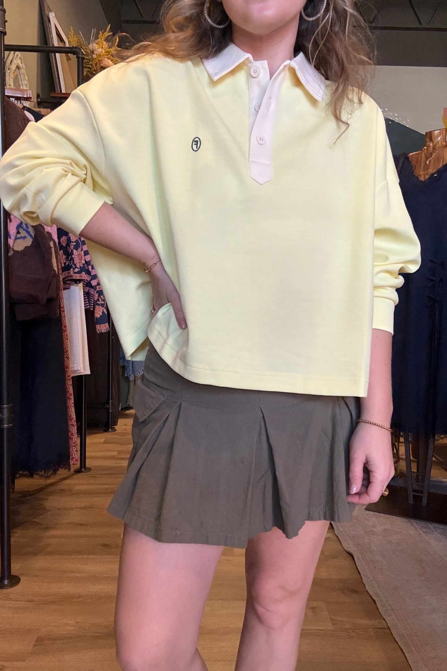 A person wearing a pale yellow long-sleeve polo shirt with a white collar and a pleated olive green mini skirt, standing inside a clothing store.