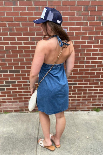 model is wearing a navy trucker hat, and a backless halter denim mini dress and carrying a straw handbag