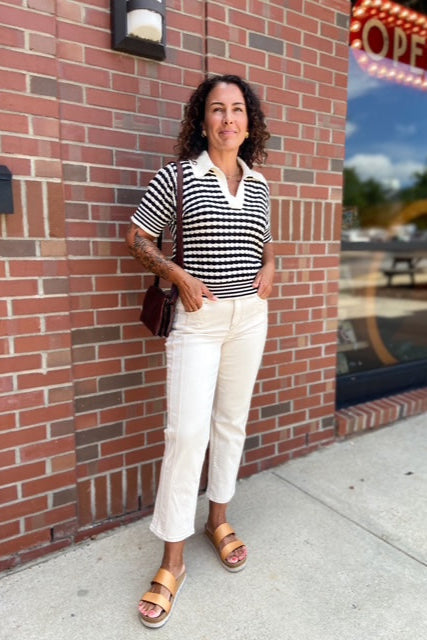 Woman in a striped shirt and white pants standing against a brick wall.