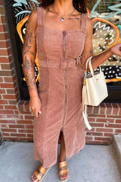 Person wearing a brown corduroy dress with tattoos visible on arms, standing against a colorful wall.
