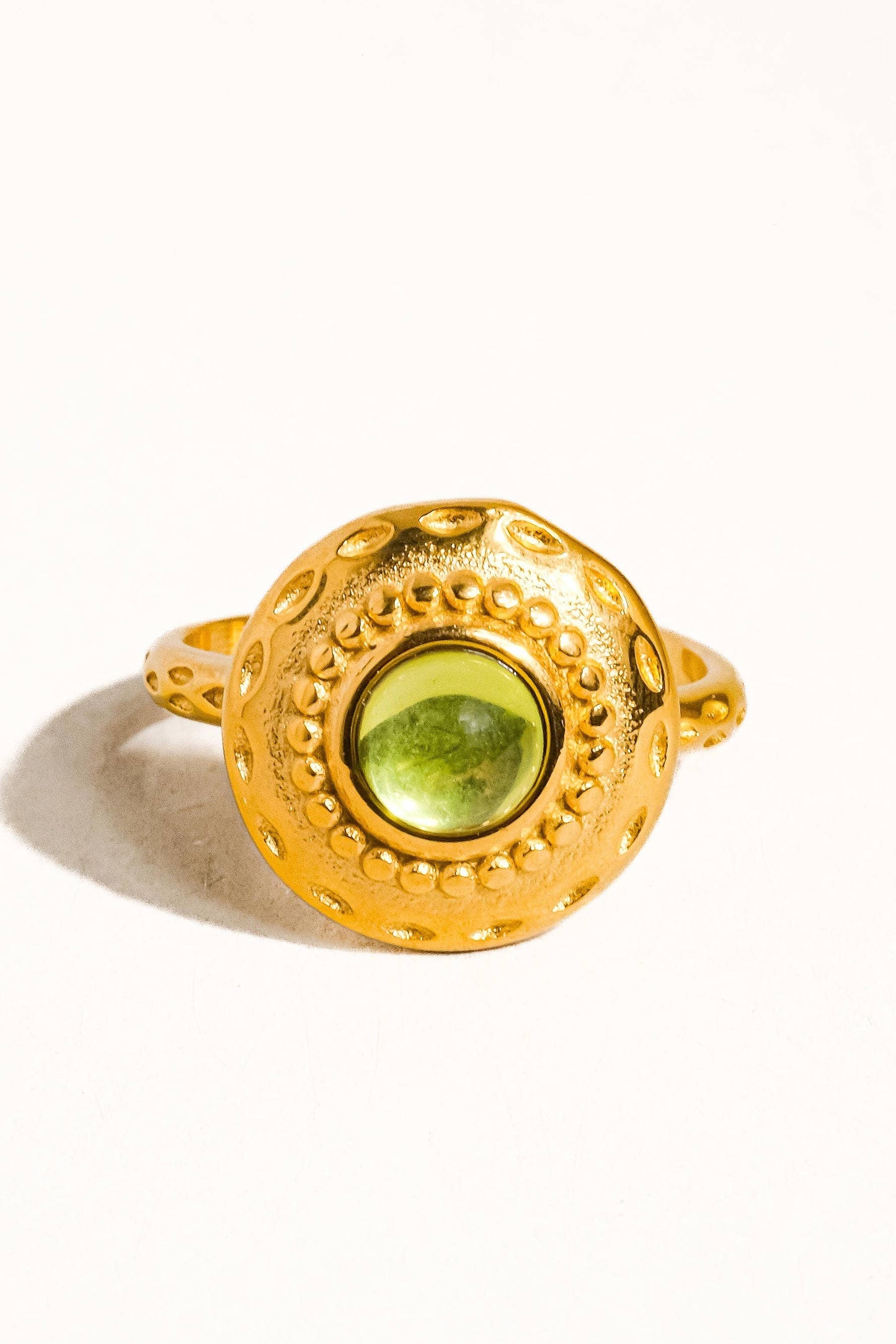 A gold ring with a large, round, textured bezel-set green gemstone, photographed on a clean white background with a soft shadow.