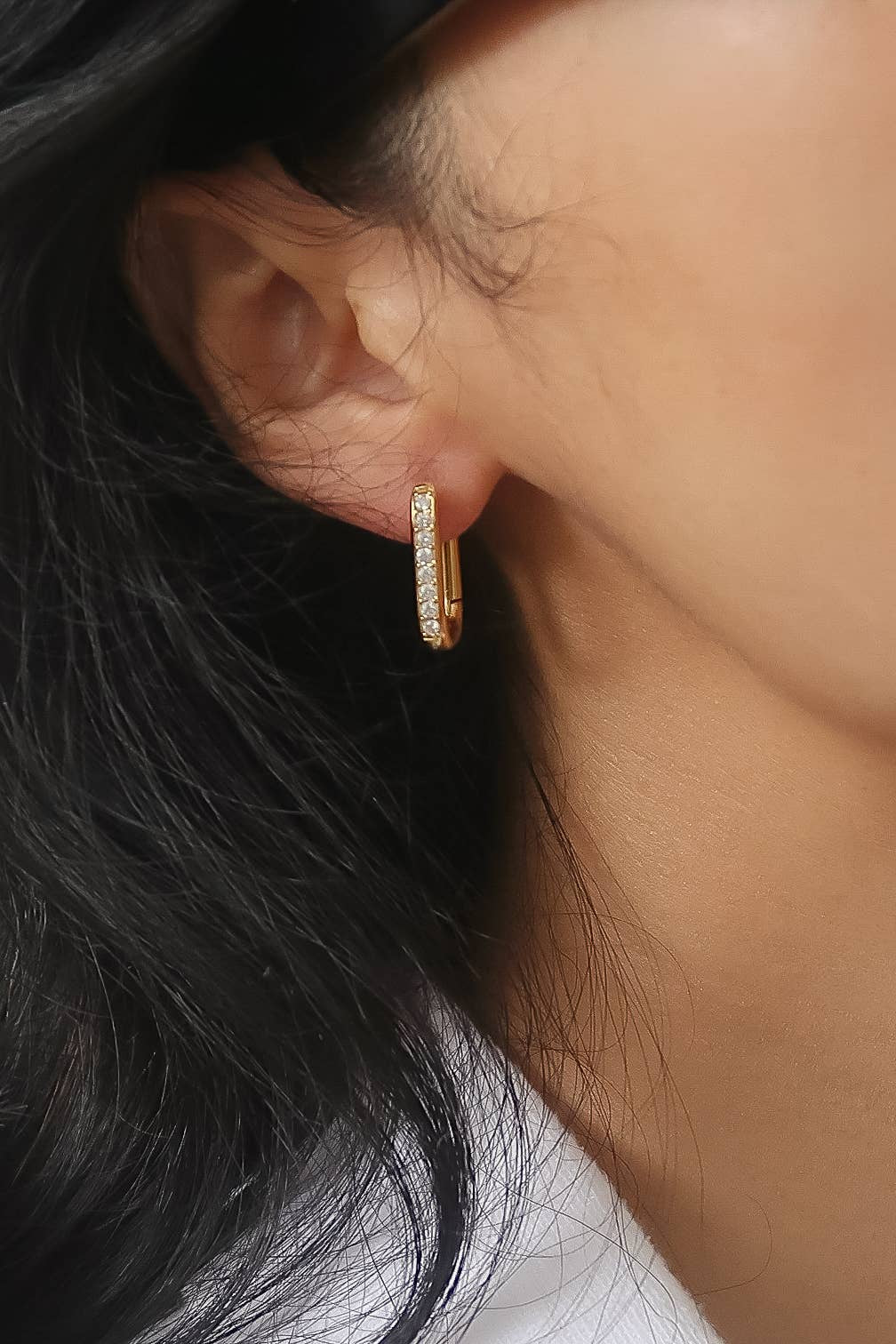 A close-up of a model's ear wearing a small gold huggie hoop earring adorned with clear pavé stones.