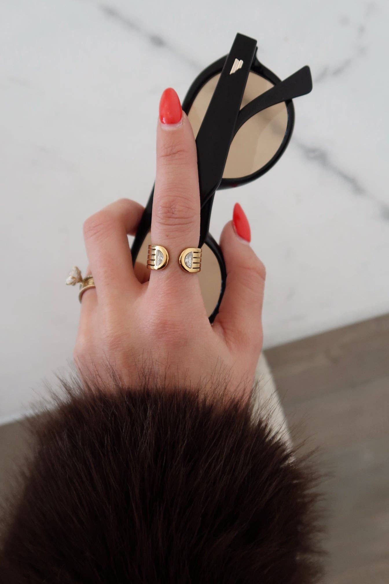 Hand wearing gold rings holding a black sandal with a white sole on a light wooden floor.