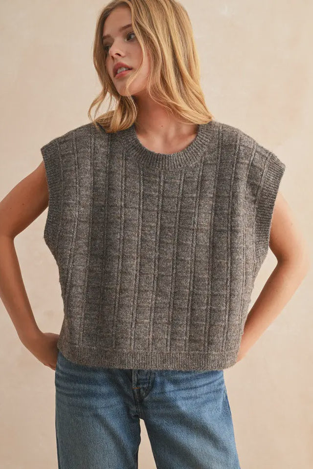 Woman wearing a gray sleeveless knitted top and blue jeans against a beige background