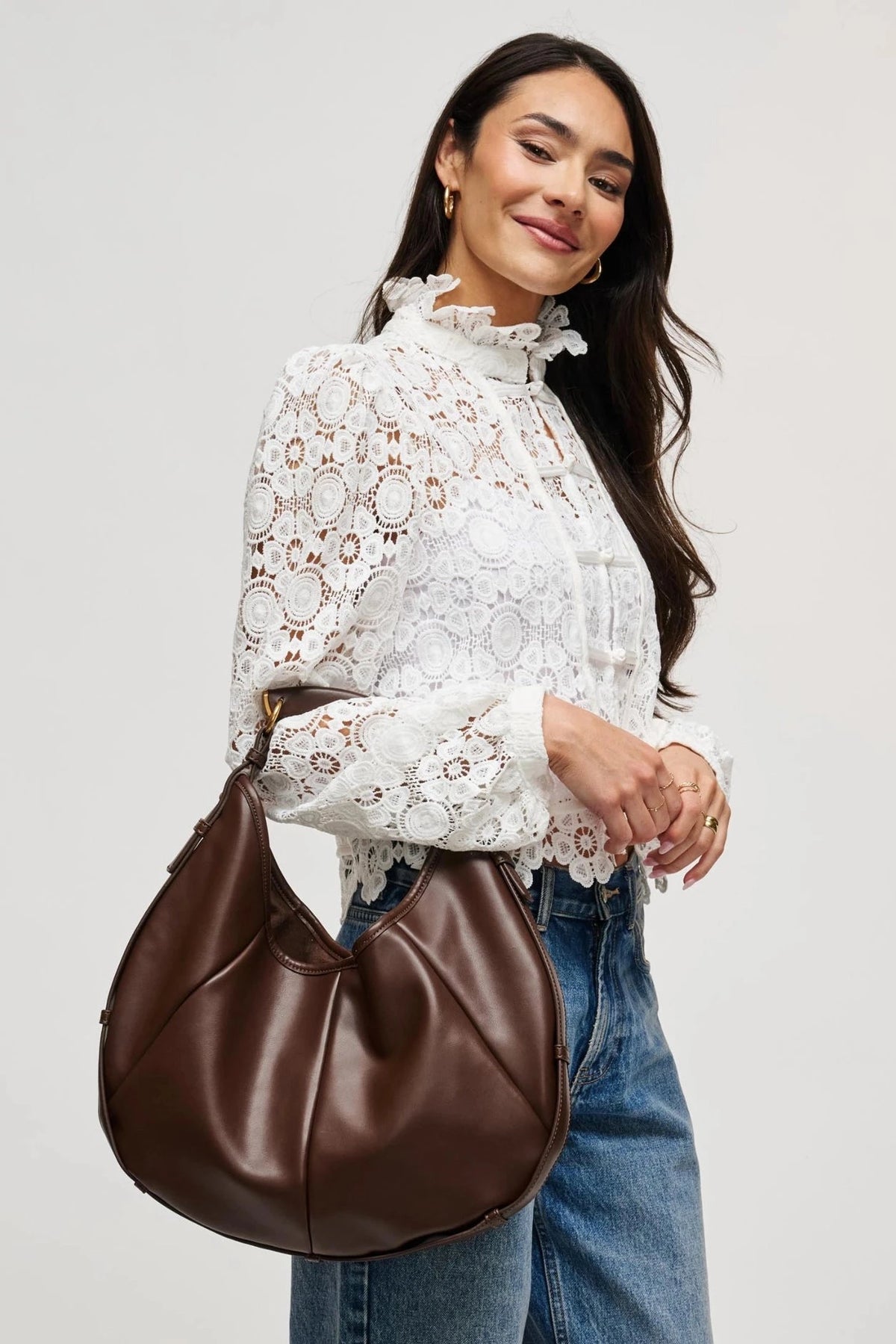 A woman wearing a white lace blouse and blue jeans carries a brown leather shoulder bag against a plain light gray background.