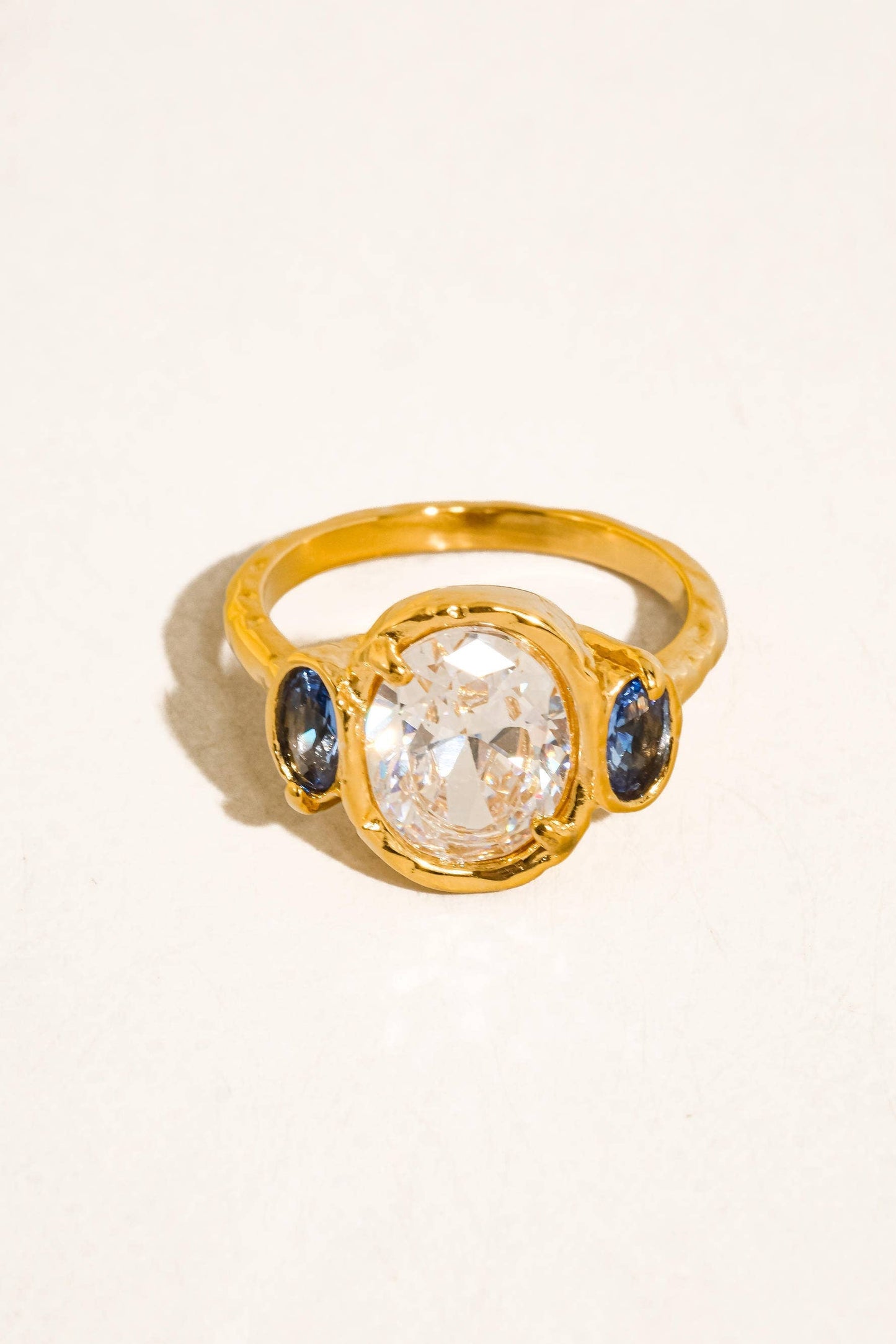 A gold ring featuring a large oval clear center stone flanked by two small round blue stones, photographed on a plain light background.
