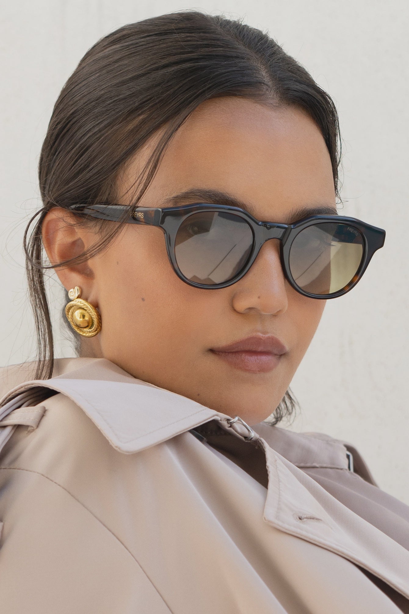 Woman wearing sunglasses and a beige trench coat against a light background