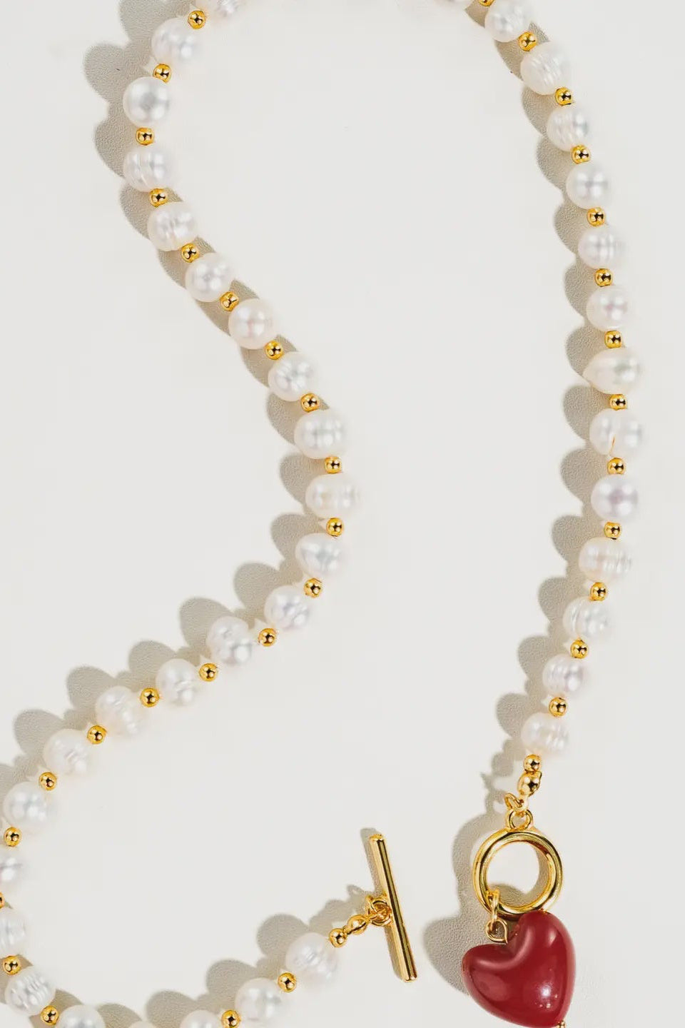 Pearl necklace with gold accents and a red heart charm on a white background