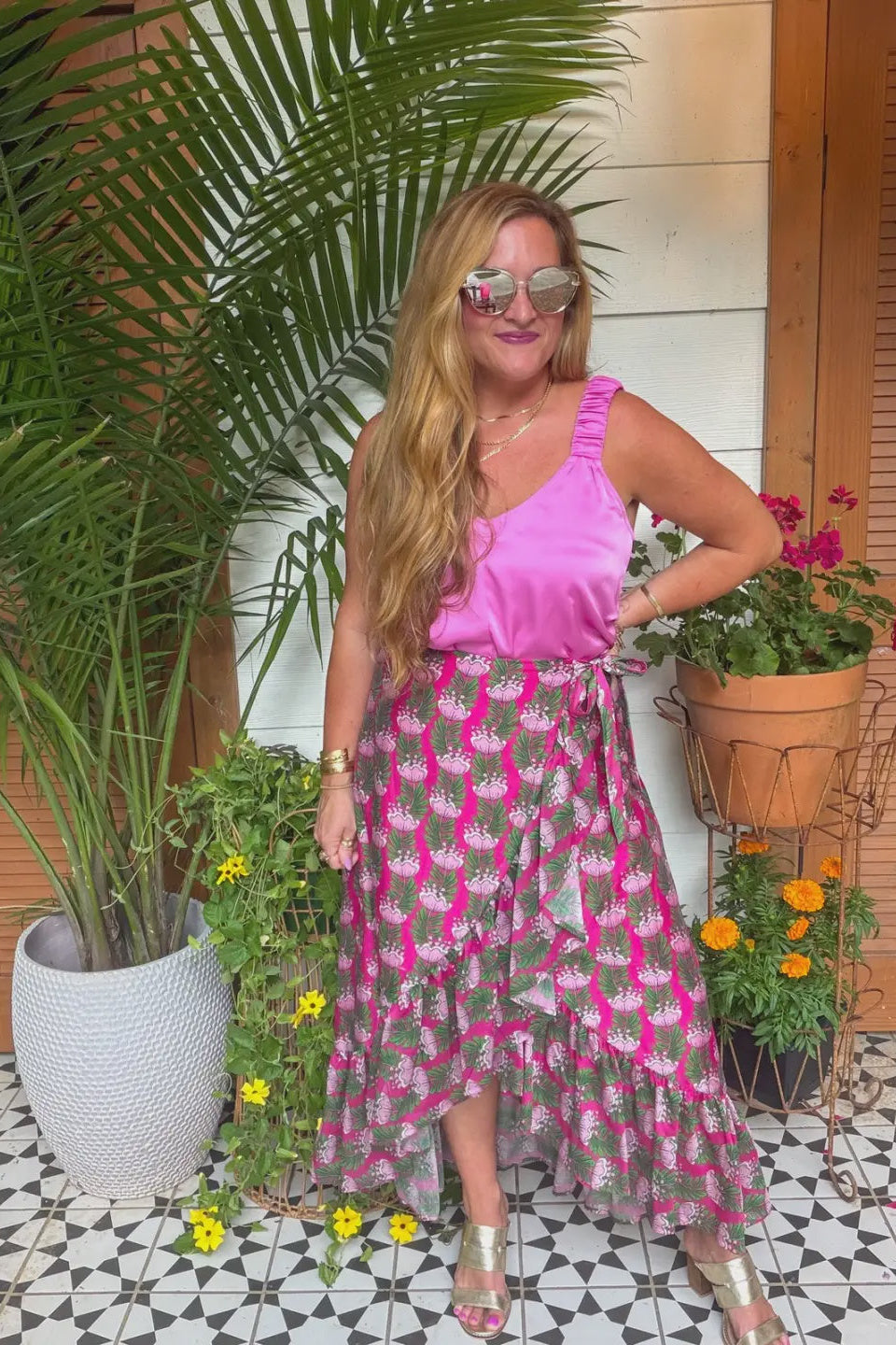 A woman modeling a two-piece outfit consisting of a bright pink ruffled sleeveless top and a matching high-low midi skirt with a pink and gray tropical floral print, styled with gold metallic sandals and sunglasses in an outdoor patio setting with potted plants.