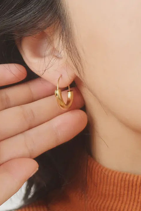 A person showing a single gold-colored U-shape hoop earring in their ear.