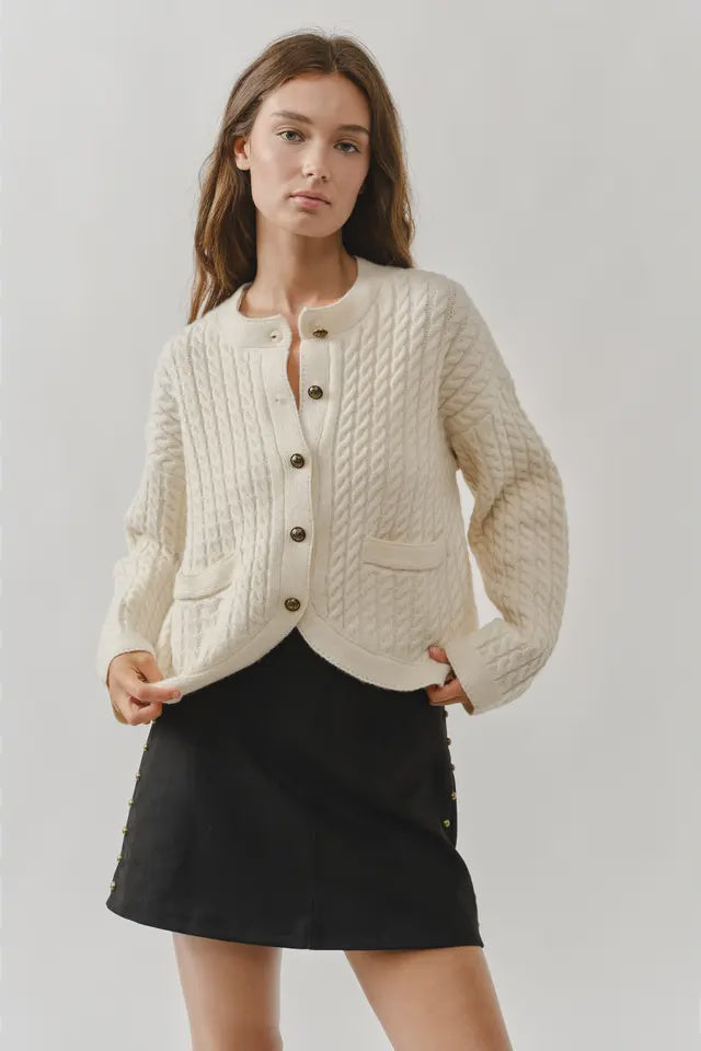 Woman wearing a cream knitted cardigan over a black skirt on a plain background