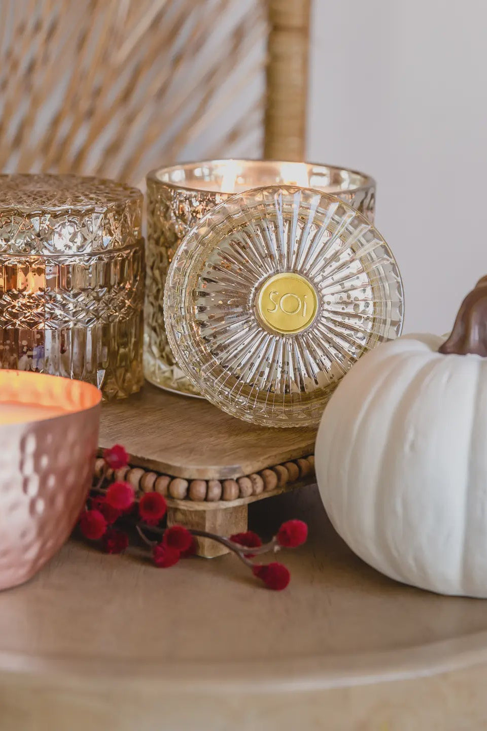 A small, square-shaped pumpkin-scented candle with a shimmering, metallic copper-colored lid, placed on a wooden surface alongside a larger, round candle. In the background, there is a pumpkin and some decorative red berries.