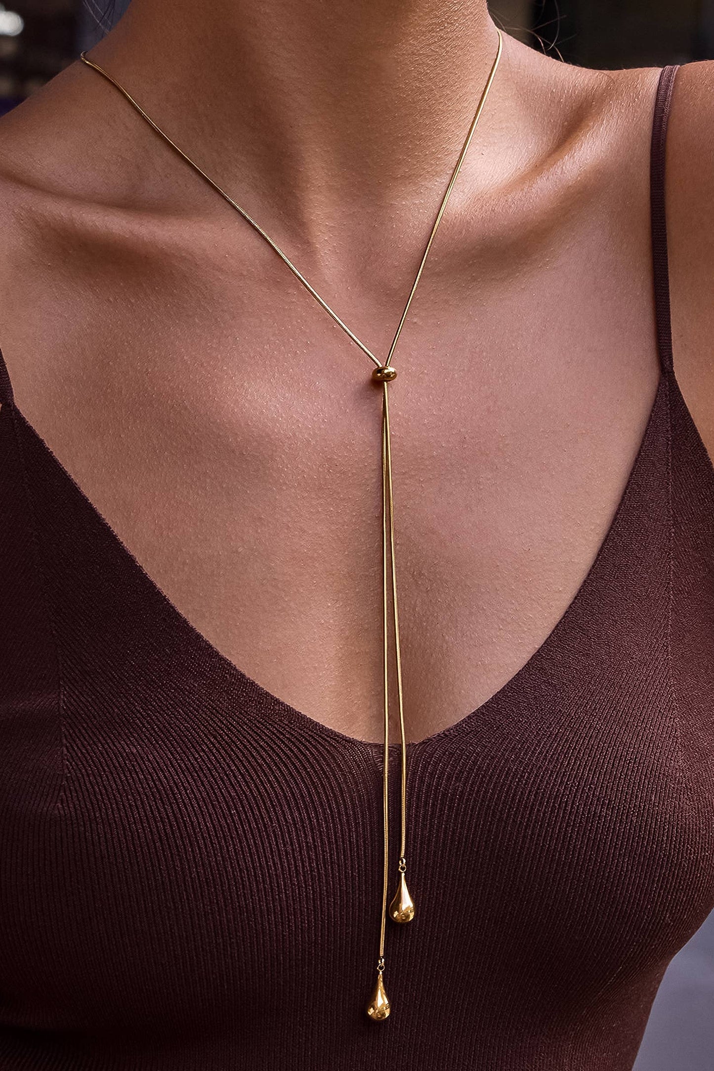 A close-up of a model wearing a delicate gold Y-necklace with two long drop chains ending in small teardrop beads, styled over a dark brown ribbed camisole top.