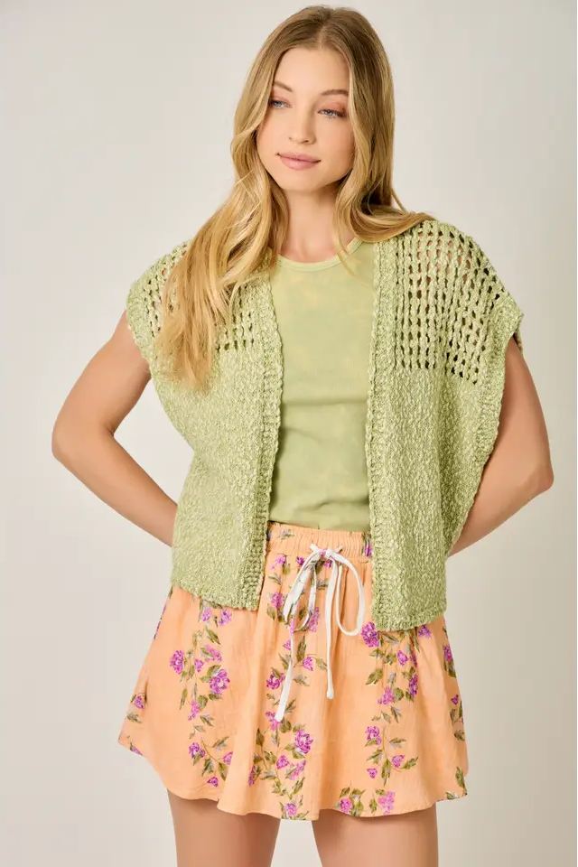 Woman wearing a green knitted vest over a light green top and floral skirt on a plain background