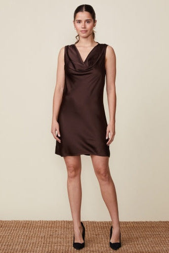 A woman modeling a sleeveless cowl-neck mini dress in dark brown satin, standing against a plain beige studio wall on a woven jute floor.