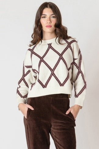 Woman wearing a cream sweater with red geometric patterns and brown pants on a white background