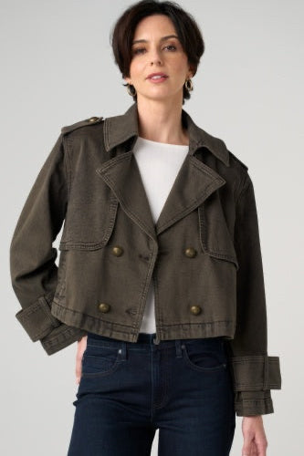 A woman wearing a cropped double-breasted utility jacket in dark olive green, styled with a white top and dark blue jeans, against a light gray studio background.